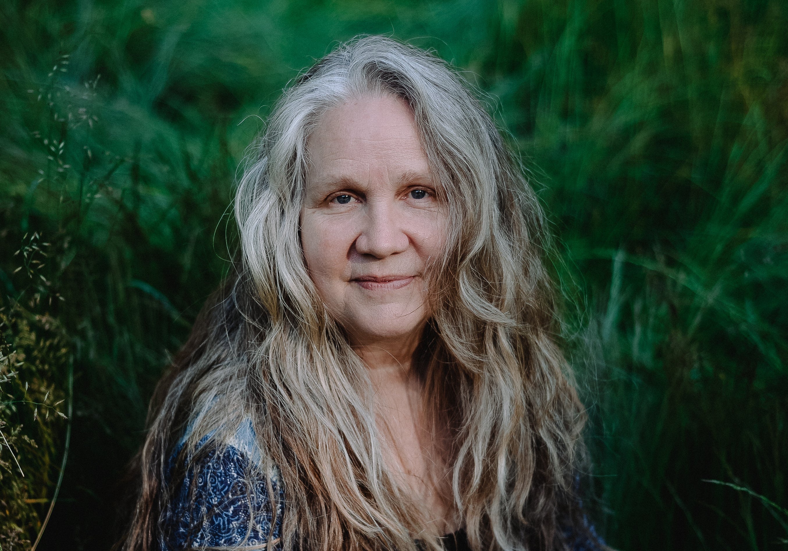 A woman with long, wavy gray hair sits in a grassy outdoor setting, wearing a blue floral print dress and holding a thin stick.