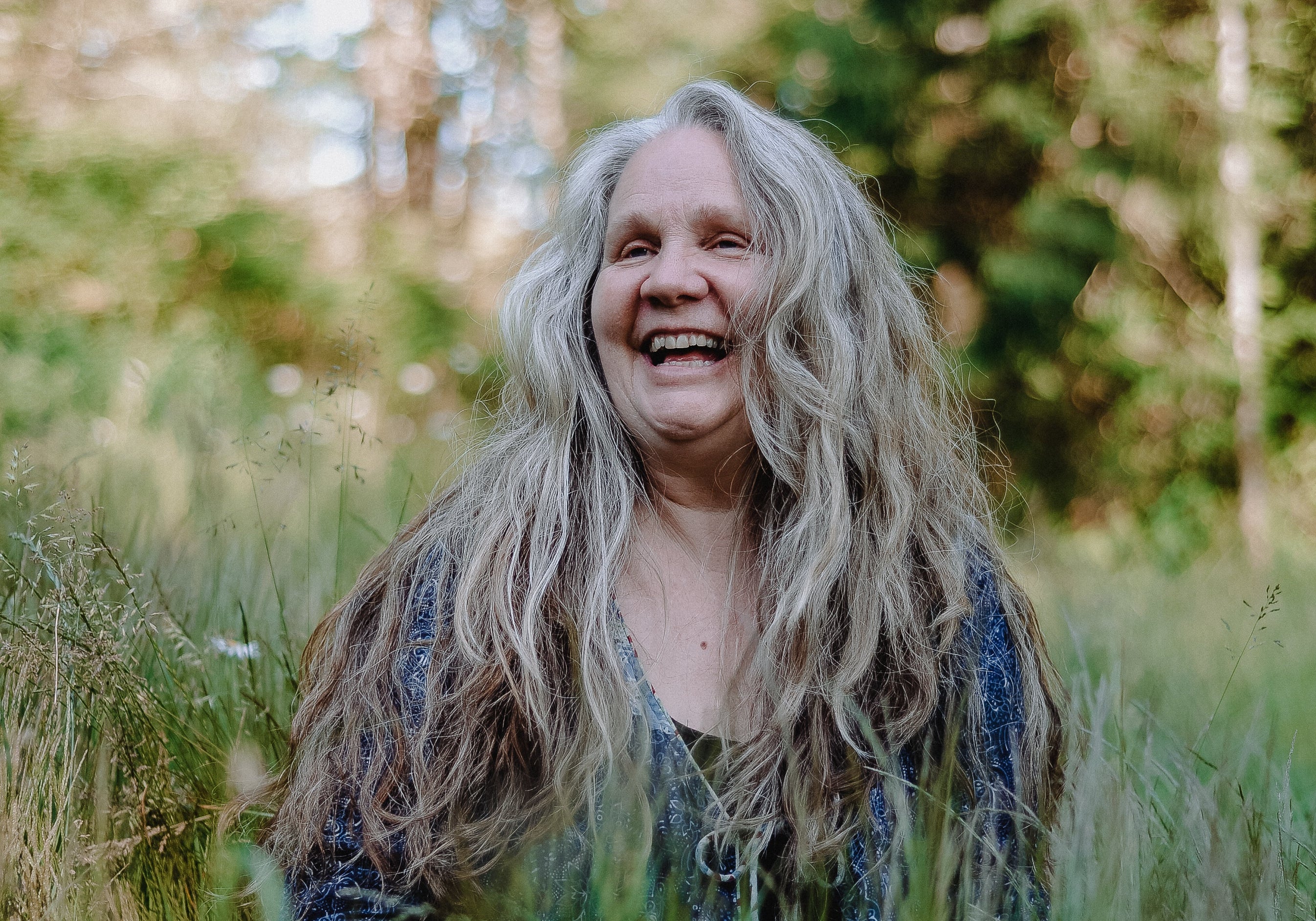 A smiling woman with long, messy gray hair stands in a grassy outdoor setting with trees in the background.