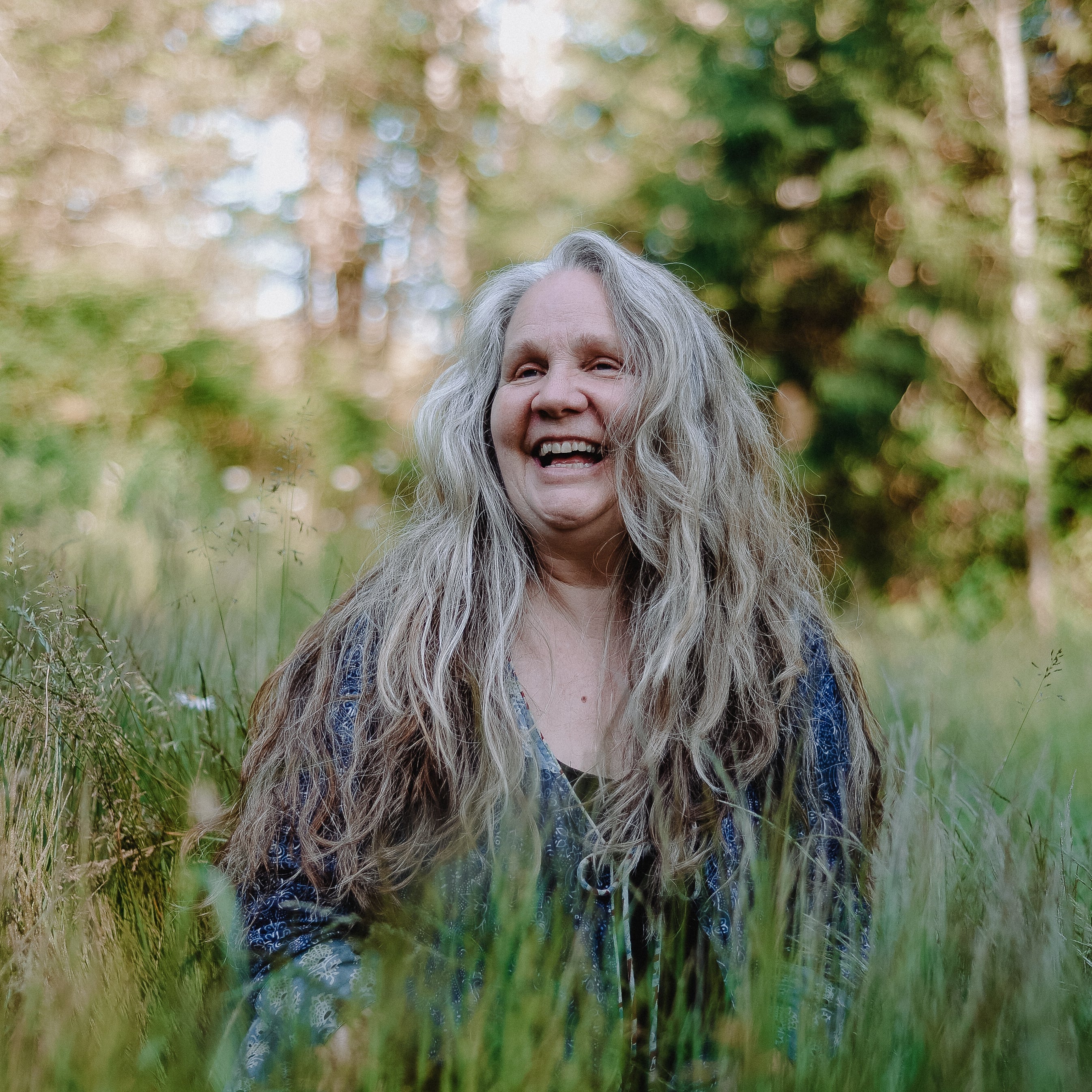 A smiling woman with long, messy gray hair stands in a grassy outdoor setting with trees in the background.