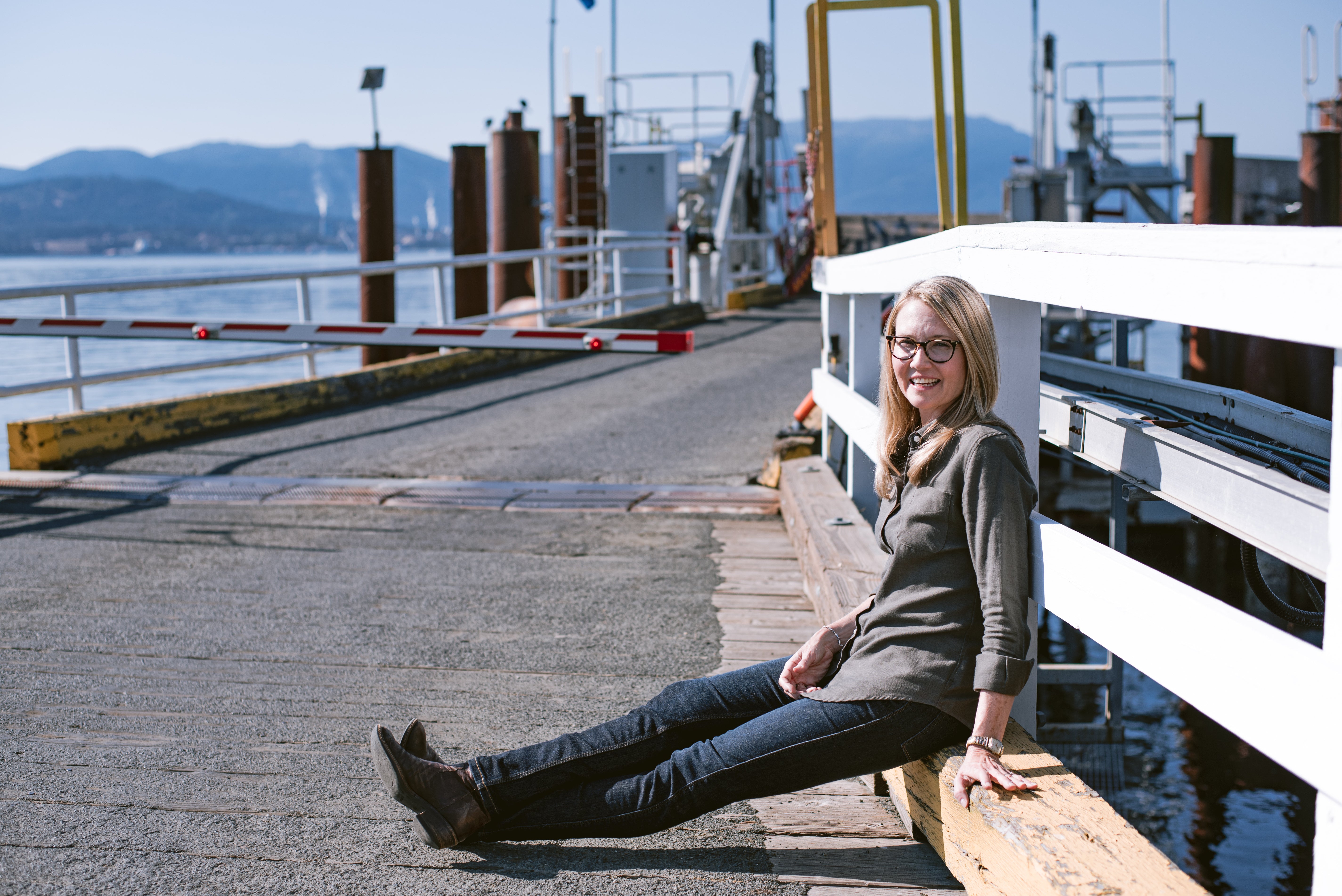Woman in casual sweater and jeans smiling while sitting on a dockside railing