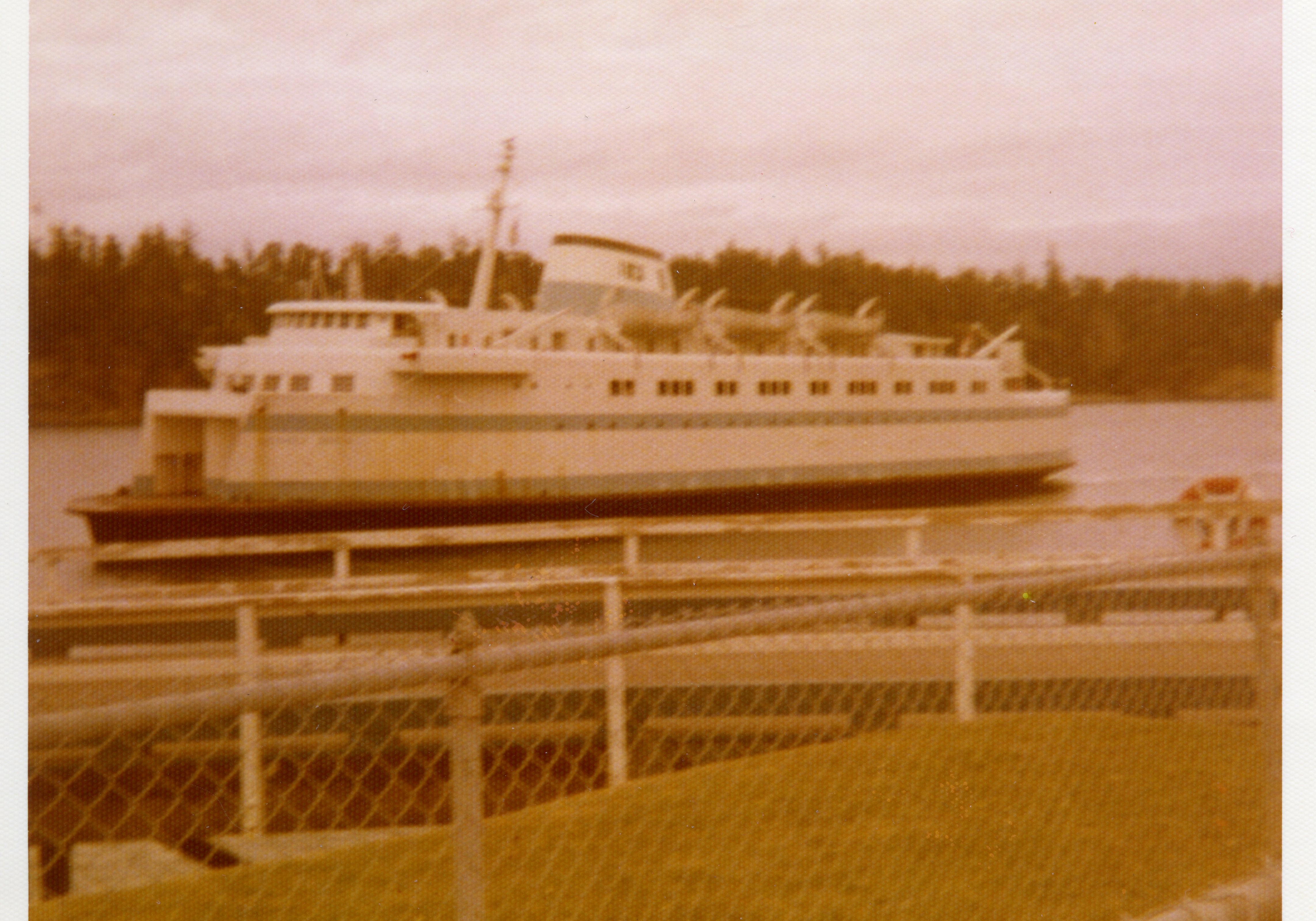 Vintage-style photograph of a ferry boat on water with a chain-link fence in the foreground.