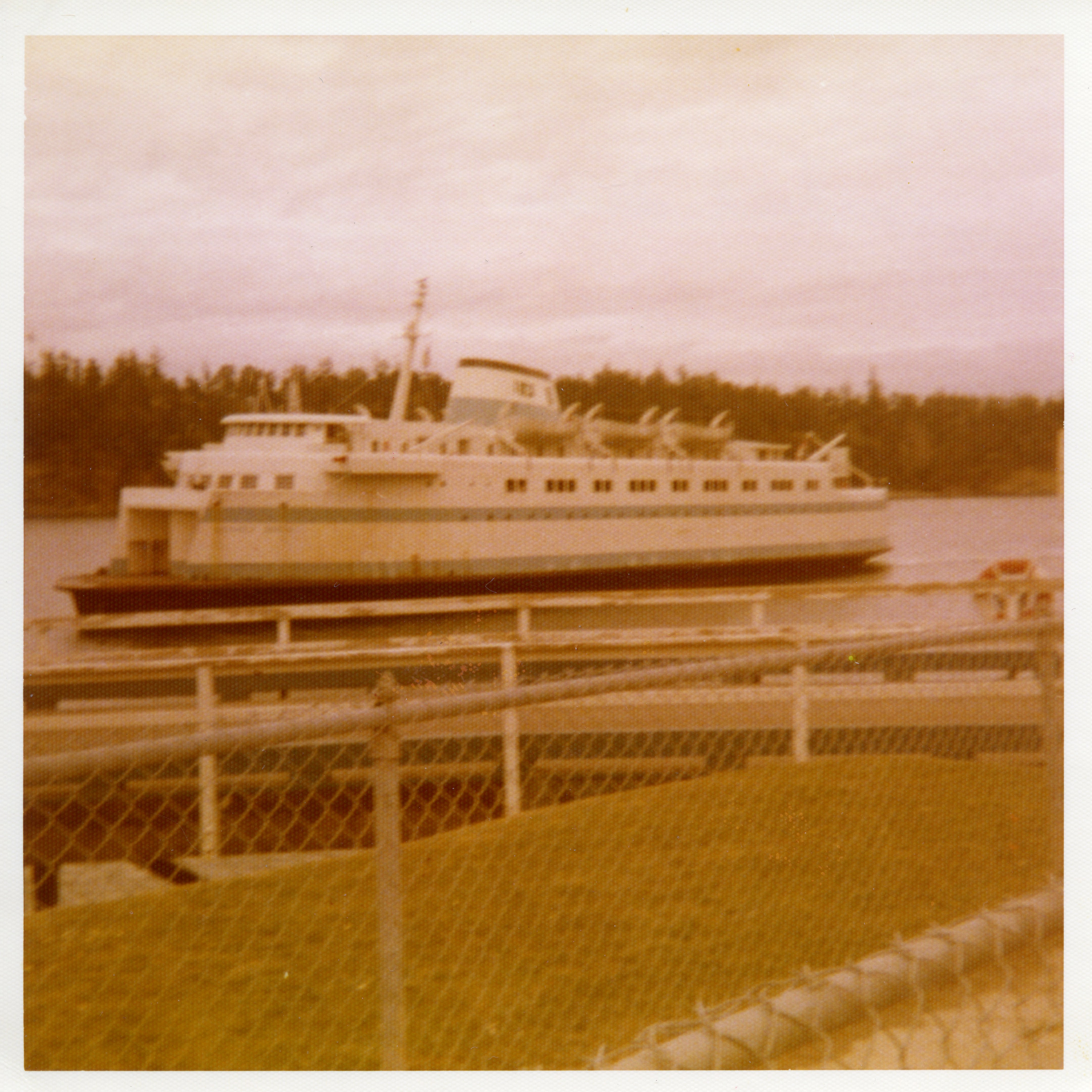 Vintage-style photograph of a ferry boat on water with a chain-link fence in the foreground.
