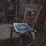 A rustic wooden chair in a garden setting with a magazine titled 'FOLKLIFE' resting on the seat, alongside a metal galvanized milk canister. In the background, a person is partially visible behind a wire mesh screen door.