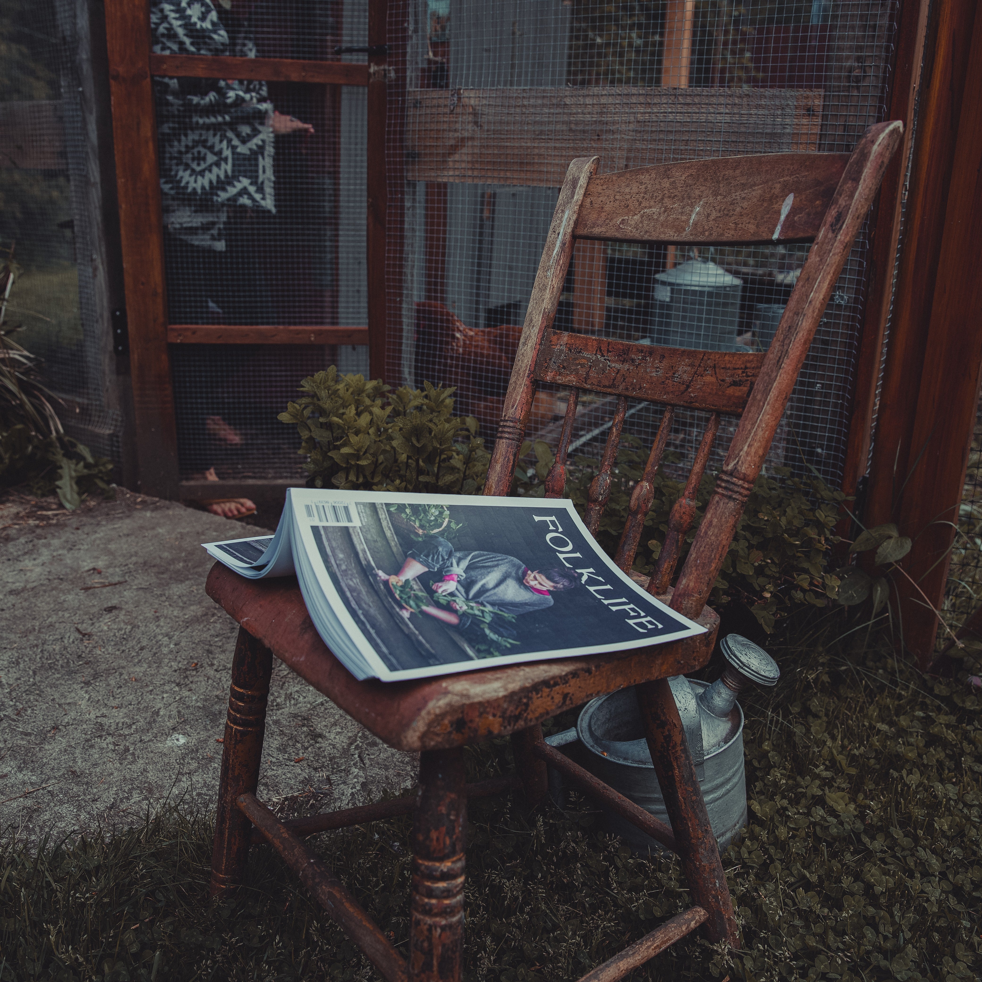 A rustic wooden chair in a garden setting with a magazine titled 'FOLKLIFE' resting on the seat, alongside a metal galvanized milk canister. In the background, a person is partially visible behind a wire mesh screen door.