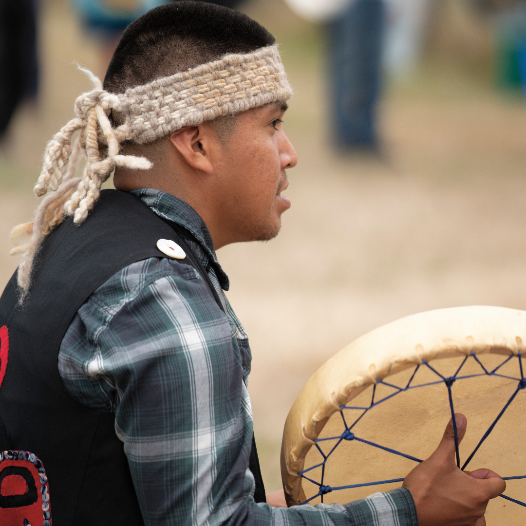 Patrick Aleck playing a hand drum while wearing a woven cedar headband