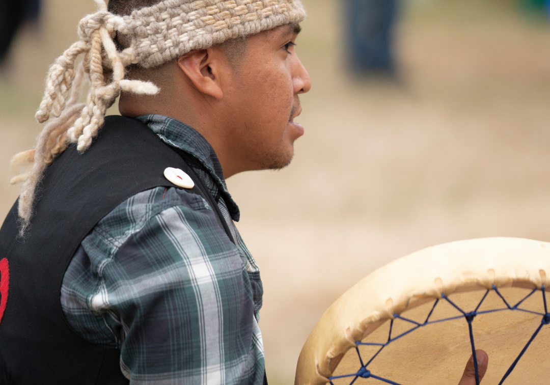 Patrick Aleck playing a hand drum while wearing a woven cedar headband