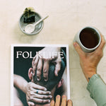 A person holds a cup of tea at the side of FOLKLIFE magazine on a light textured surface. The magazine cover shows a close-up photo of hands on a person's head, and a small bowl with a spoon and a dark wrapped item sits above the magazine.