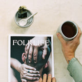 A person holds a cup of tea at the side of FOLKLIFE magazine on a light textured surface. The magazine cover shows a close-up photo of hands on a person's head, and a small bowl with a spoon and a dark wrapped item sits above the magazine.