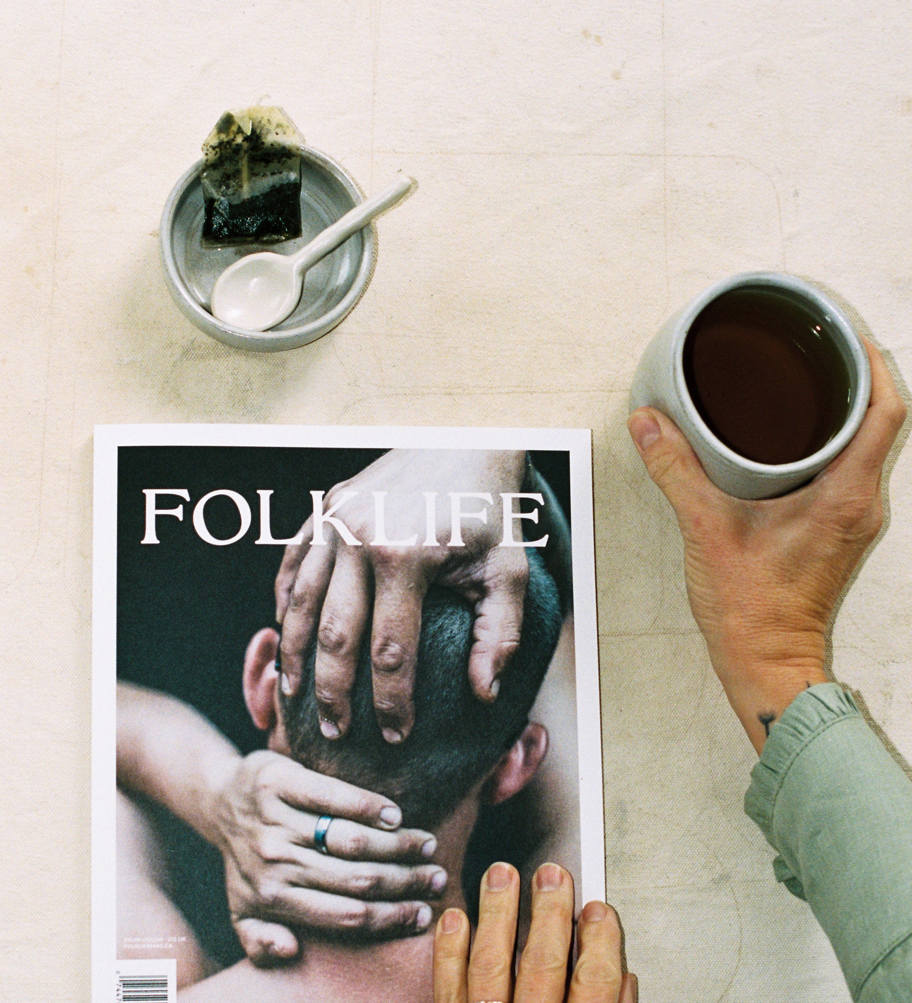 A person holds a cup of tea at the side of FOLKLIFE magazine on a light textured surface. The magazine cover shows a close-up photo of hands on a person's head, and a small bowl with a spoon and a dark wrapped item sits above the magazine.