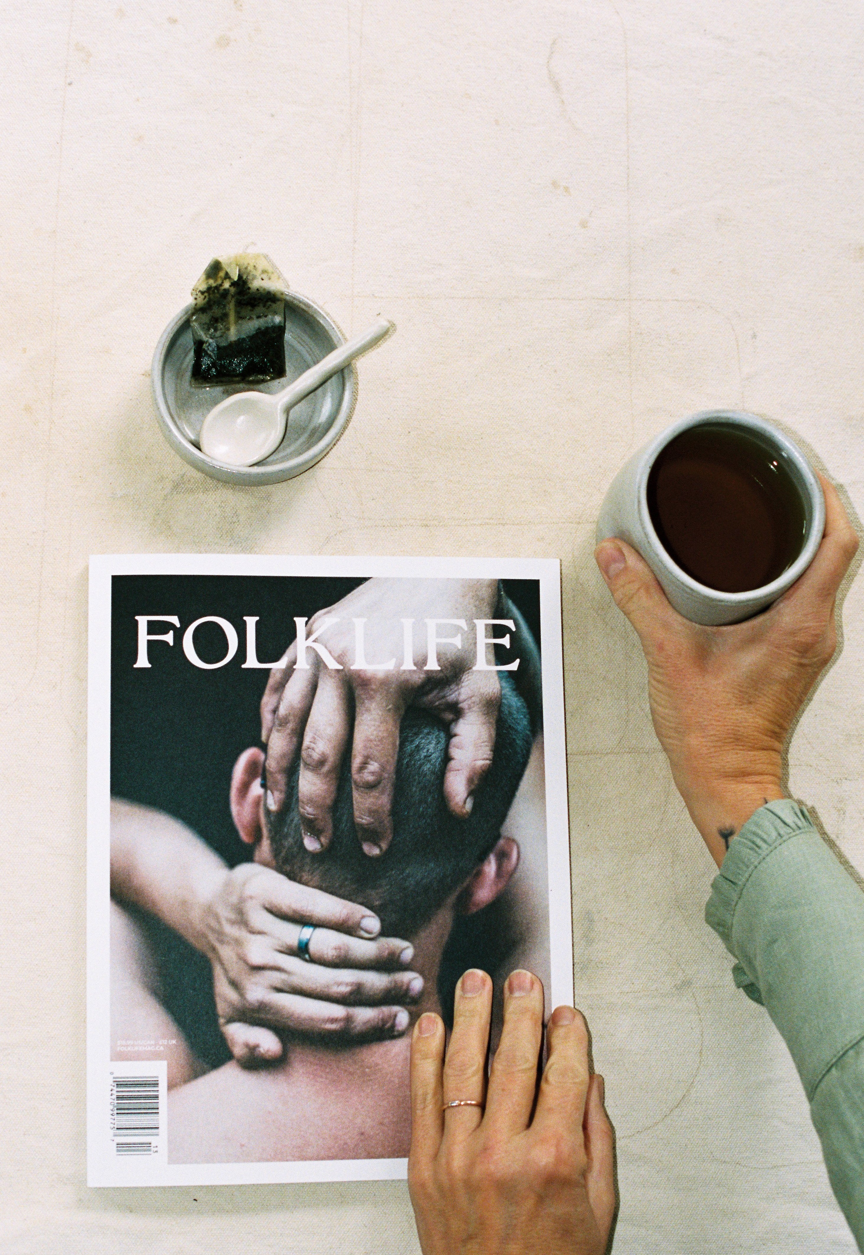 A person holds a cup of tea at the side of FOLKLIFE magazine on a light textured surface. The magazine cover shows a close-up photo of hands on a person's head, and a small bowl with a spoon and a dark wrapped item sits above the magazine.