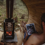 A man relaxes in a wooden cabin sauna, holding a FOLKLIFE magazine, with a black wood-burning stove and a round window showing a forest view outside.