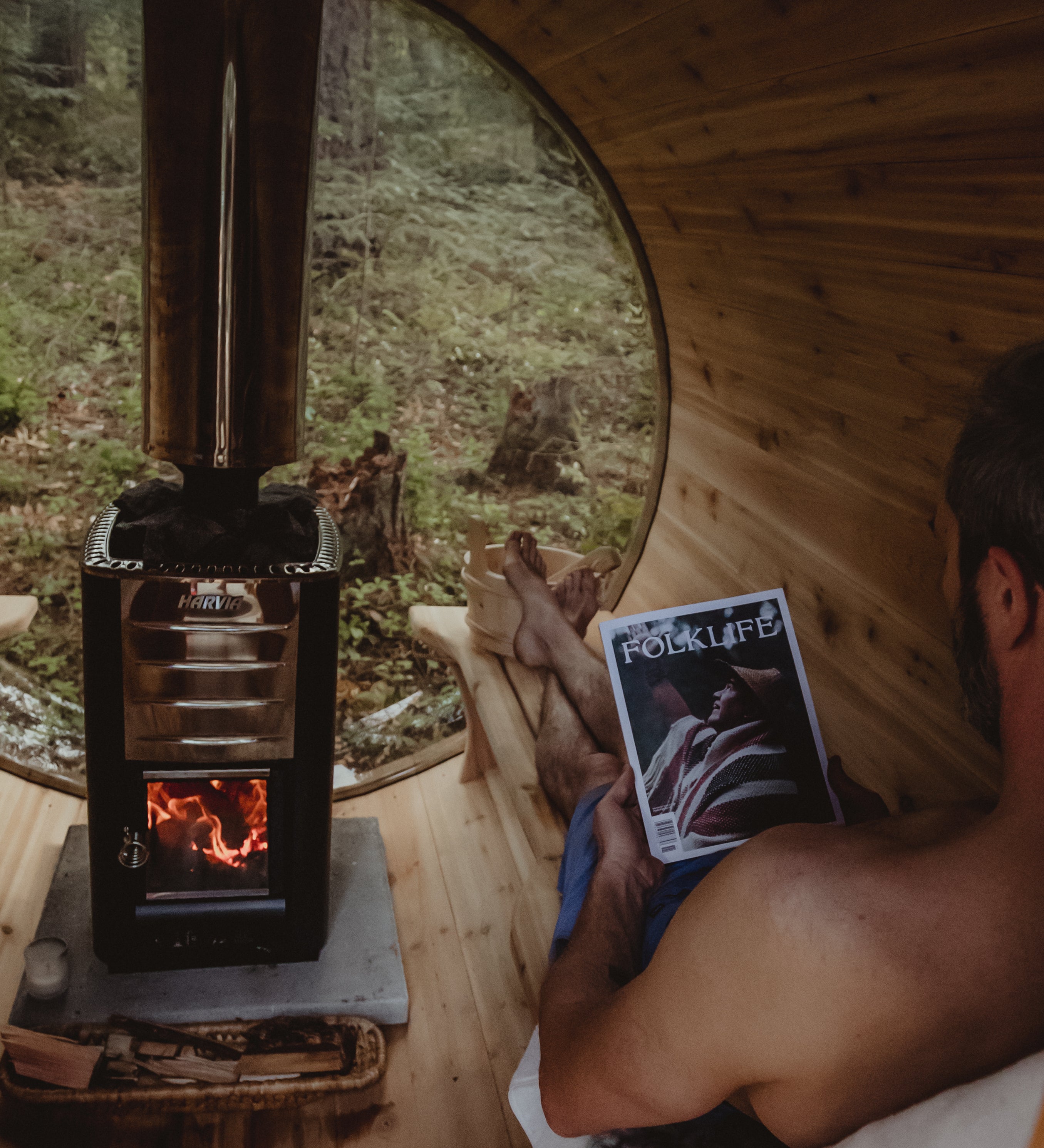 A man relaxes in a wooden cabin sauna, holding a FOLKLIFE magazine, with a black wood-burning stove and a round window showing a forest view outside.