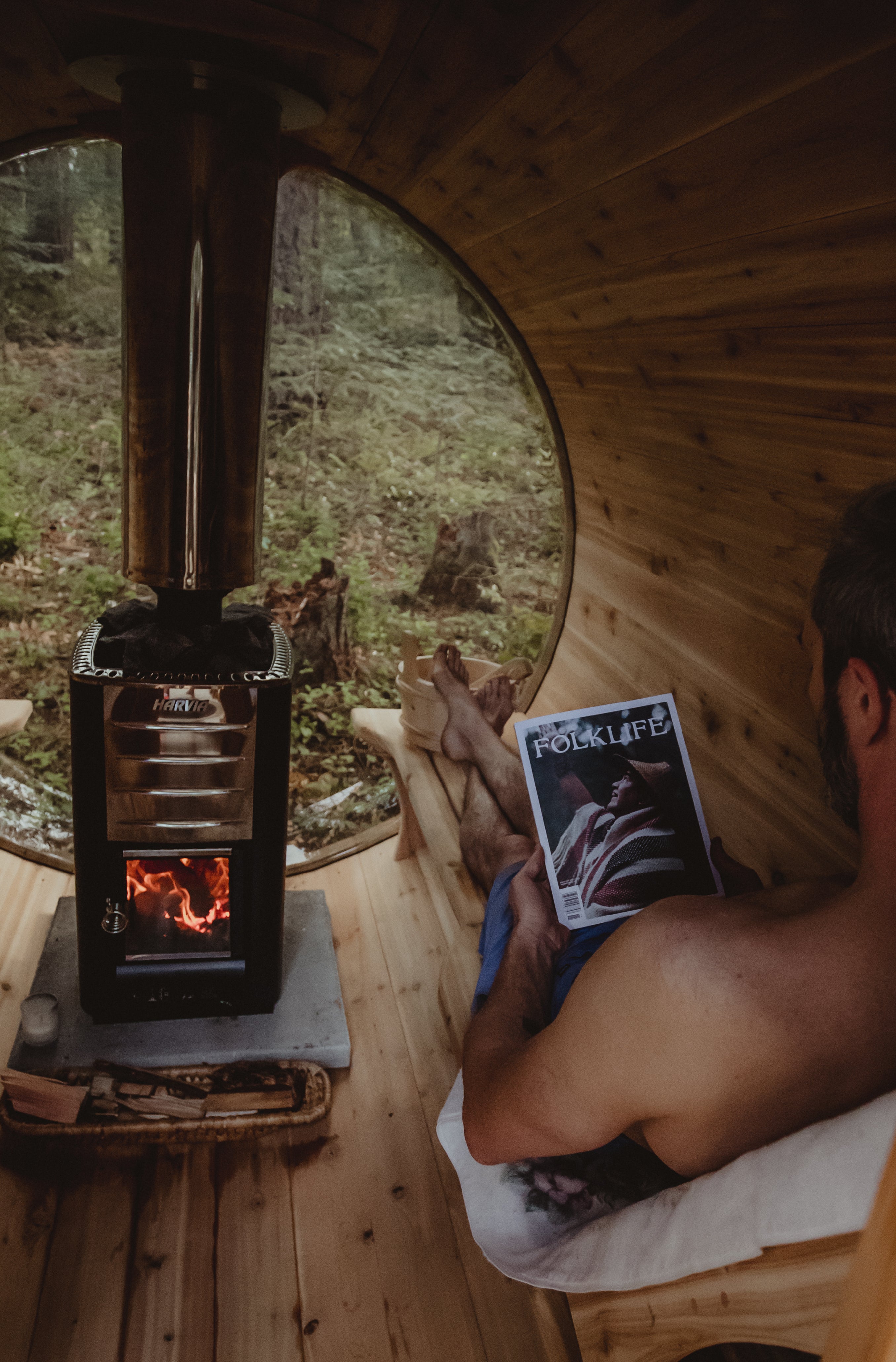 A man relaxes in a wooden cabin sauna, holding a FOLKLIFE magazine, with a black wood-burning stove and a round window showing a forest view outside.