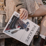 A person sitting outdoors on a wooden bench holding a FOLKLIFE magazine. The person is wearing a brown knit sweater, a leopard-print skirt, white socks, and dark brown boots, with dry leaves on the ground.