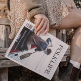 A person sitting outdoors on a wooden bench holding a FOLKLIFE magazine. The person is wearing a brown knit sweater, a leopard-print skirt, white socks, and dark brown boots, with dry leaves on the ground.
