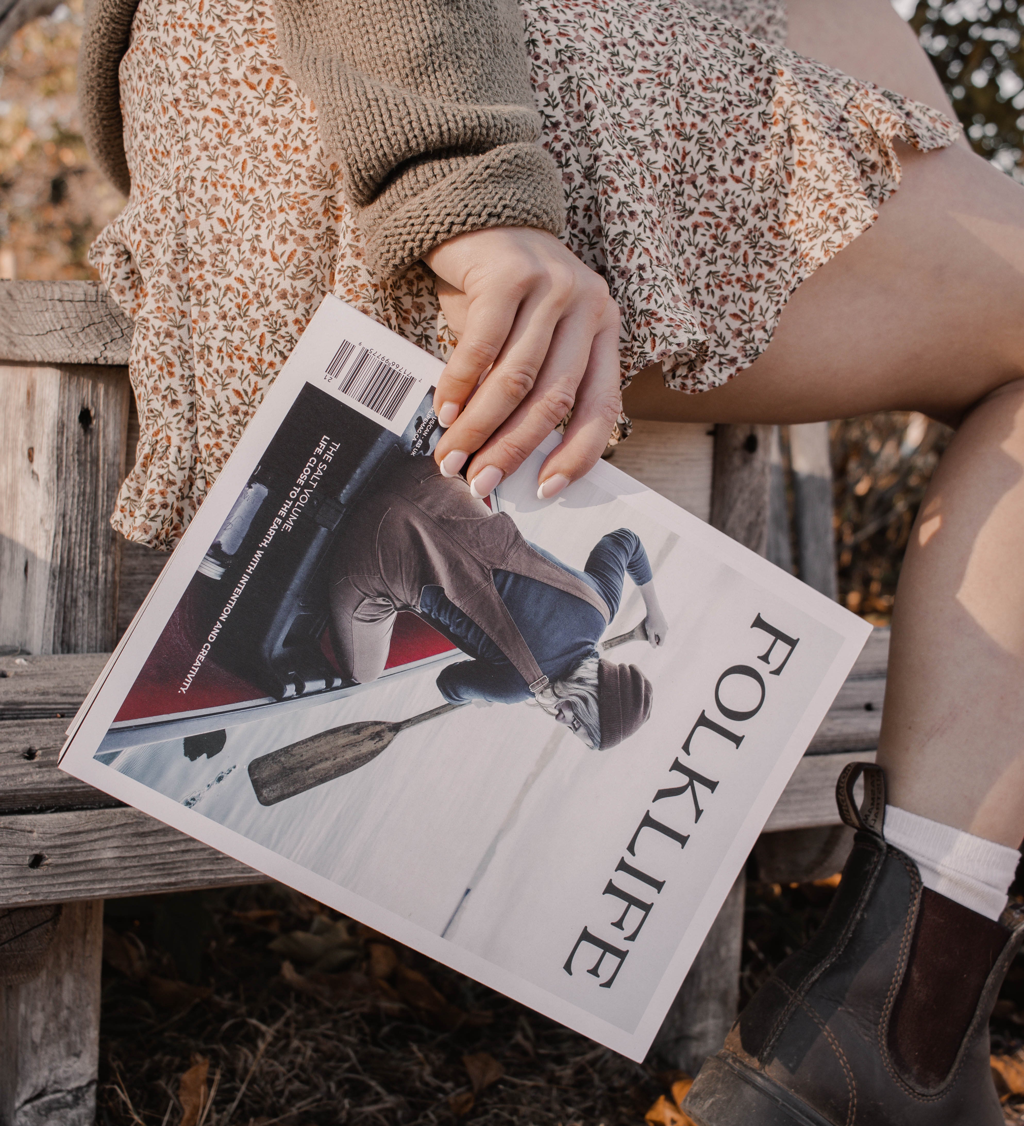 A person sitting outdoors on a wooden bench holding a FOLKLIFE magazine. The person is wearing a brown knit sweater, a leopard-print skirt, white socks, and dark brown boots, with dry leaves on the ground.