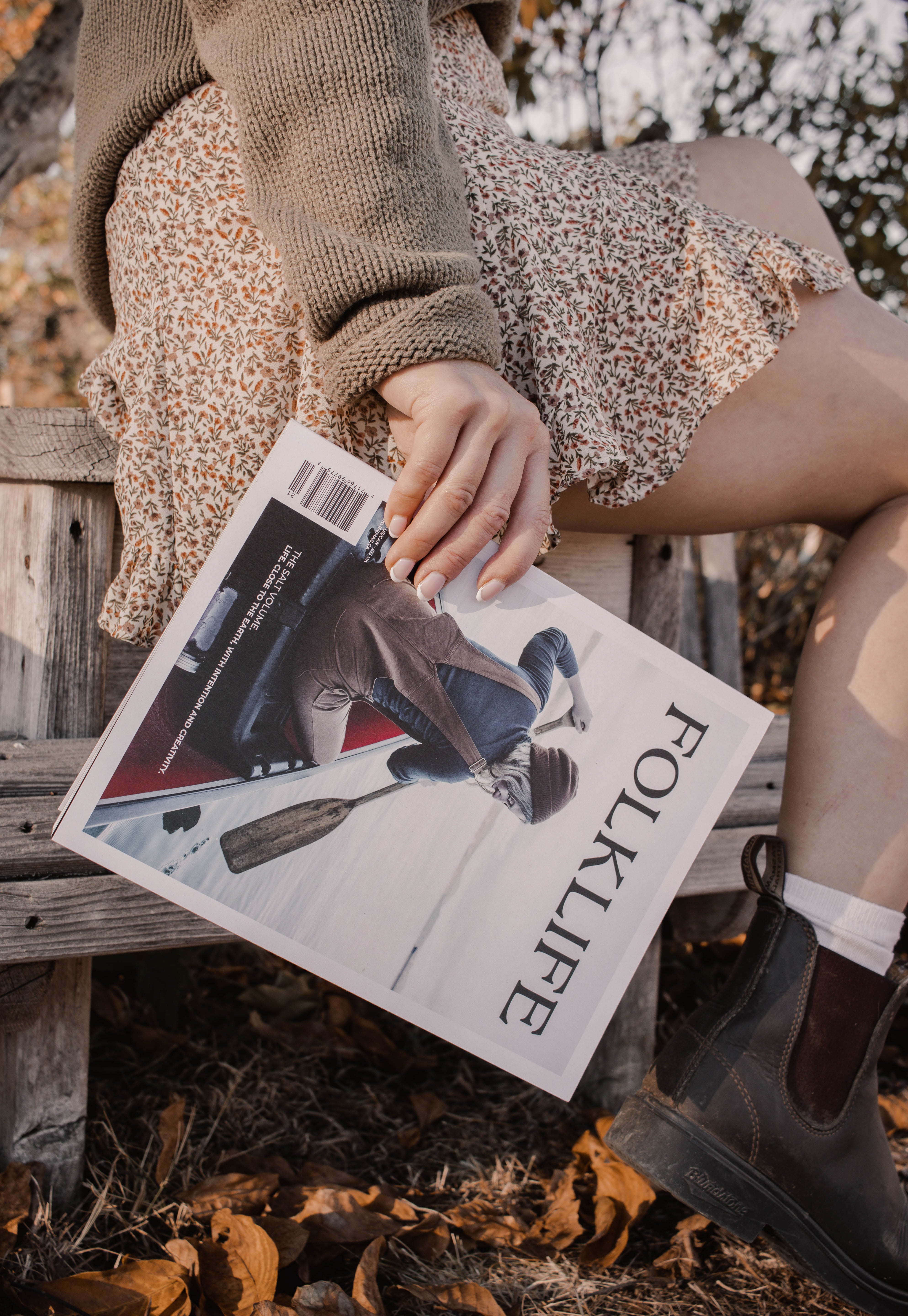 A person sitting outdoors on a wooden bench holding a FOLKLIFE magazine. The person is wearing a brown knit sweater, a leopard-print skirt, white socks, and dark brown boots, with dry leaves on the ground.