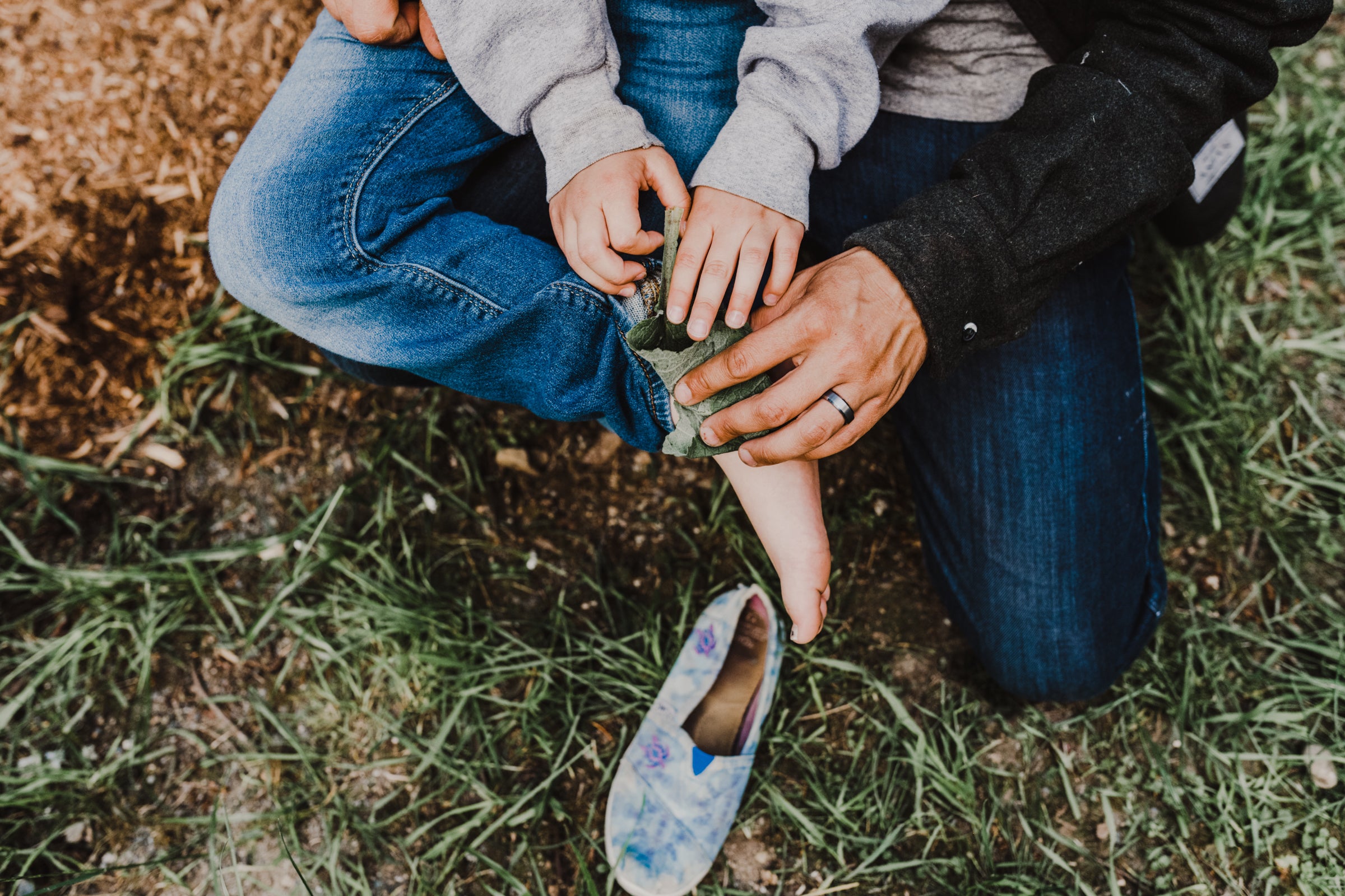 A close-up outdoor shot of a person helping a child put on a blue patterned slip-on shoe, with grass, dirt, and the adults' jeans visible.