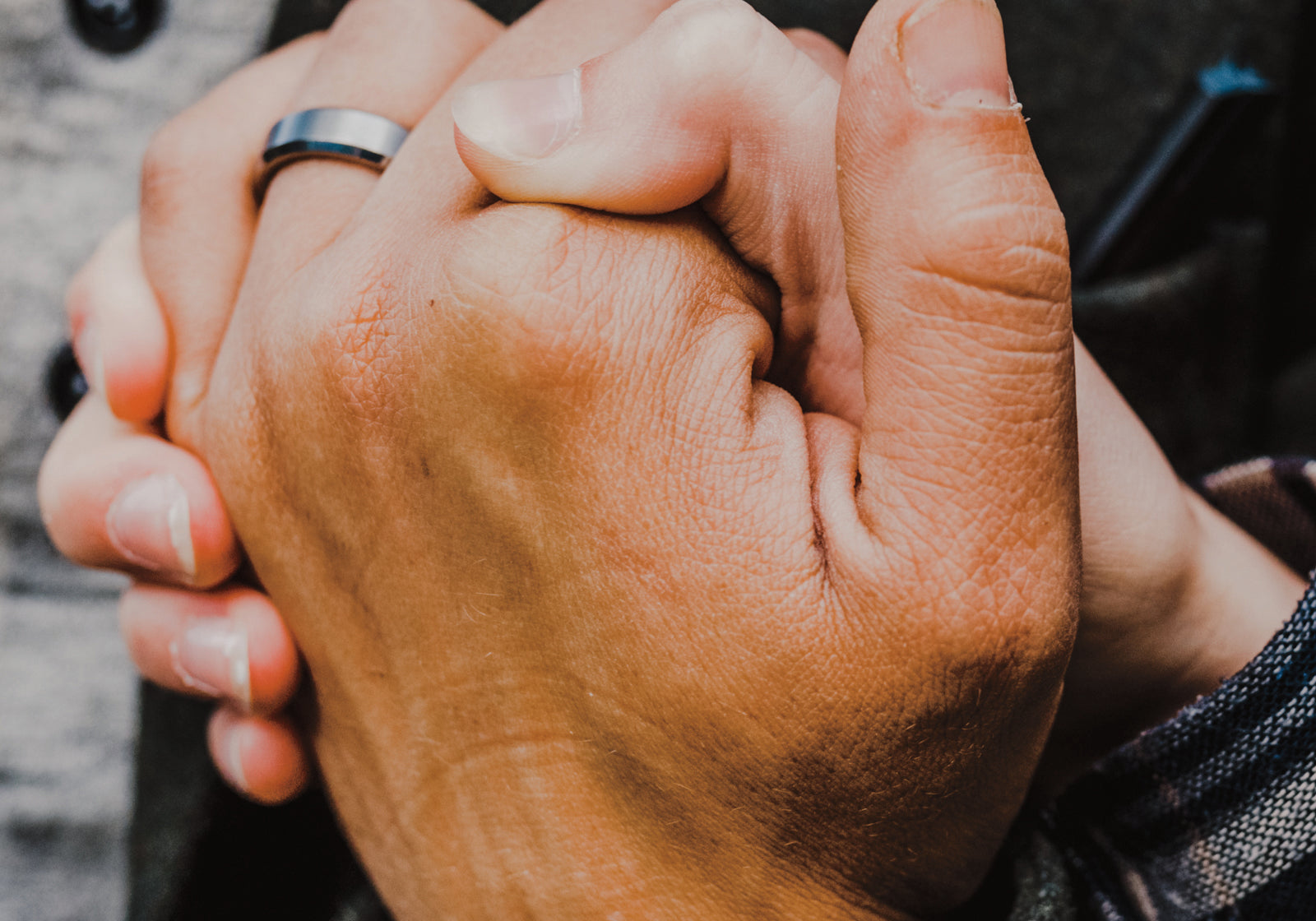 A close-up shot of two people holding hands, with one person wearing a simple silver band ring on their finger. They are dressed in dark gray and black outerwear over a gray button-up shirt.