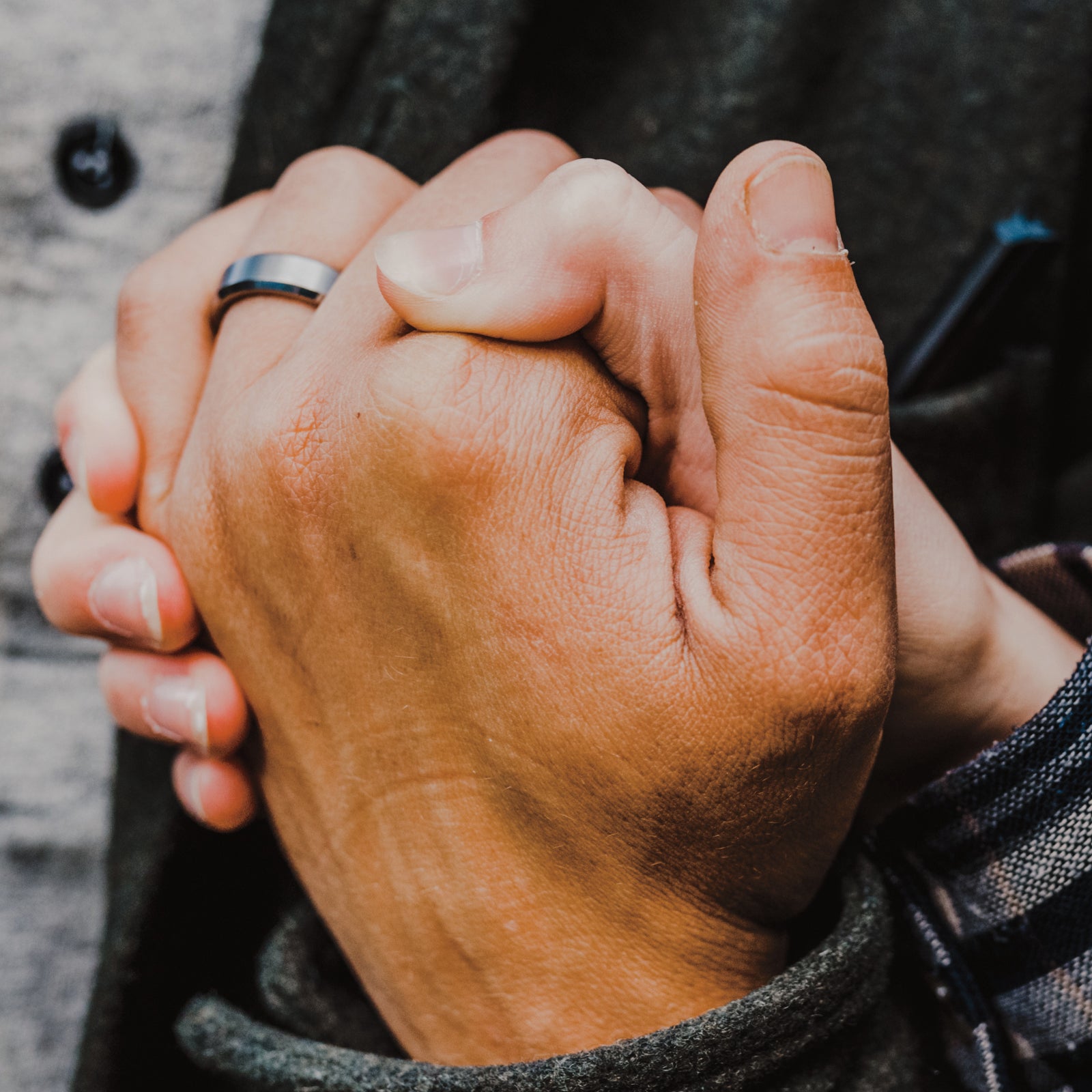A close-up shot of two people holding hands, with one person wearing a simple silver band ring on their finger. They are dressed in dark gray and black outerwear over a gray button-up shirt.