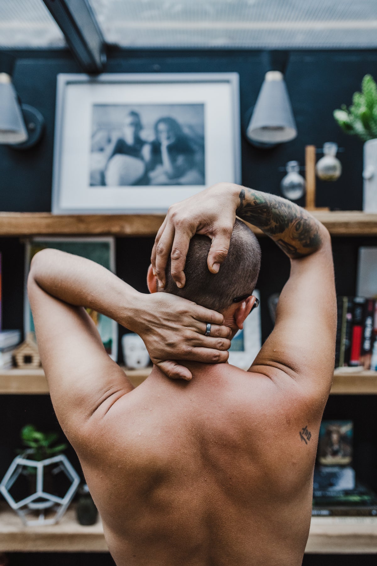 A shirtless man with tattoos on his arms is shown from behind, holding his head in pain. He is standing in a modern room with wooden shelves, books, and framed pictures in the background.
