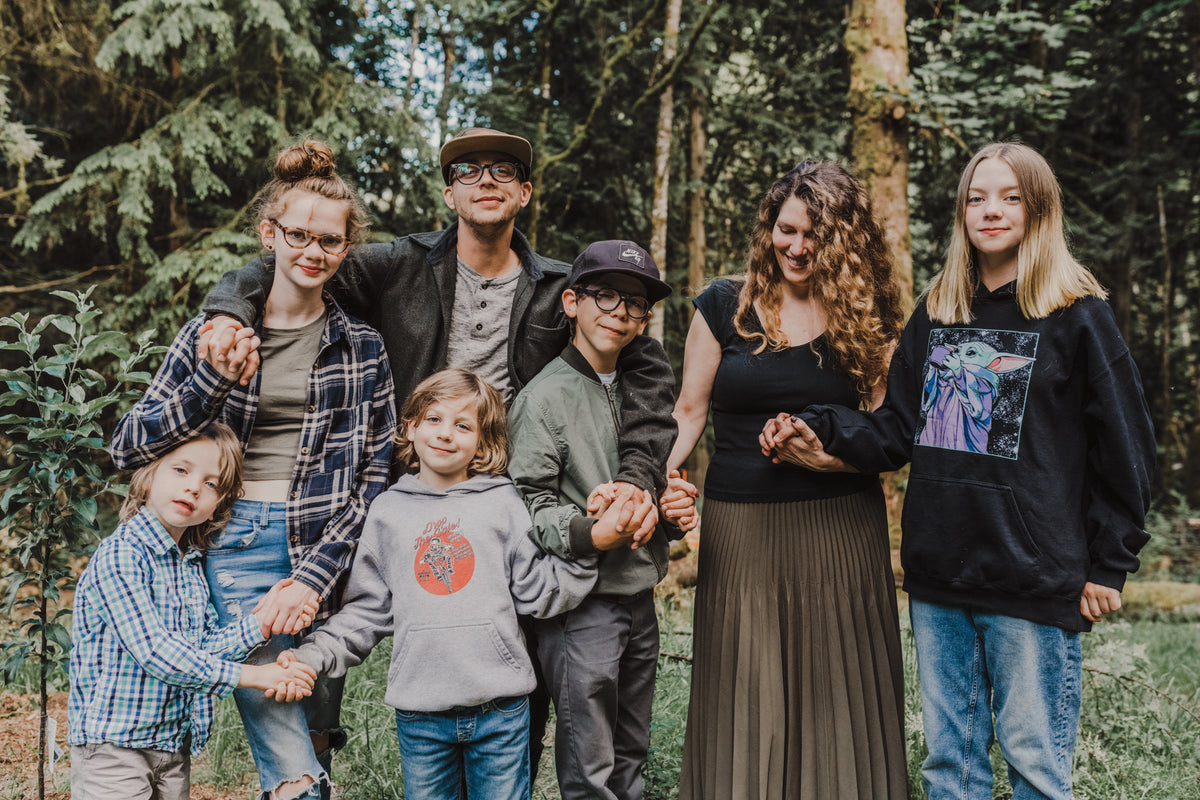 A family of seven posing together outdoors in a dense forest. The group includes a father, and six children, dressed in casual, earth-toned clothing.