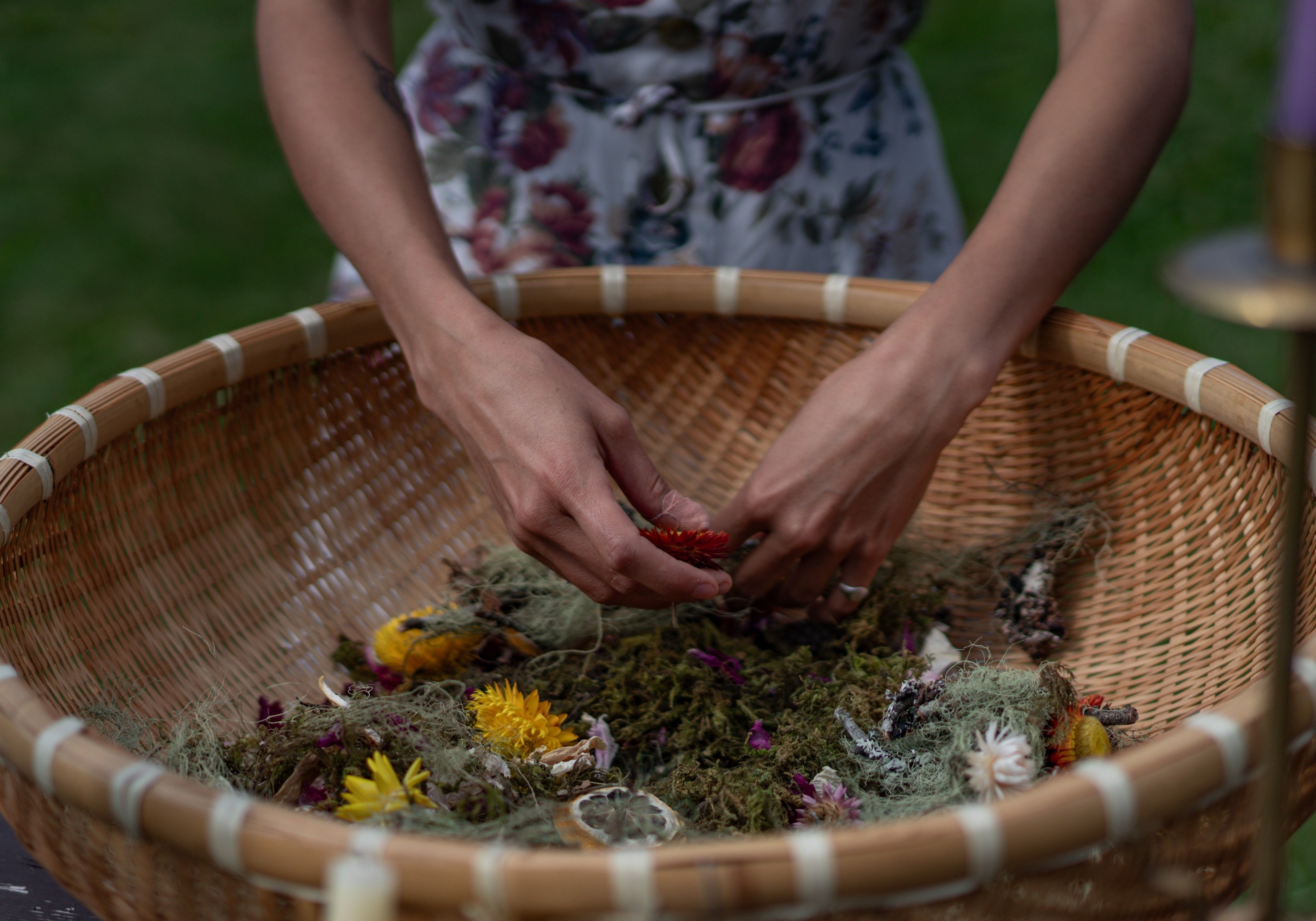 Hands arranging dried flowers and moss in woven basket centerpiece surrounded by candles