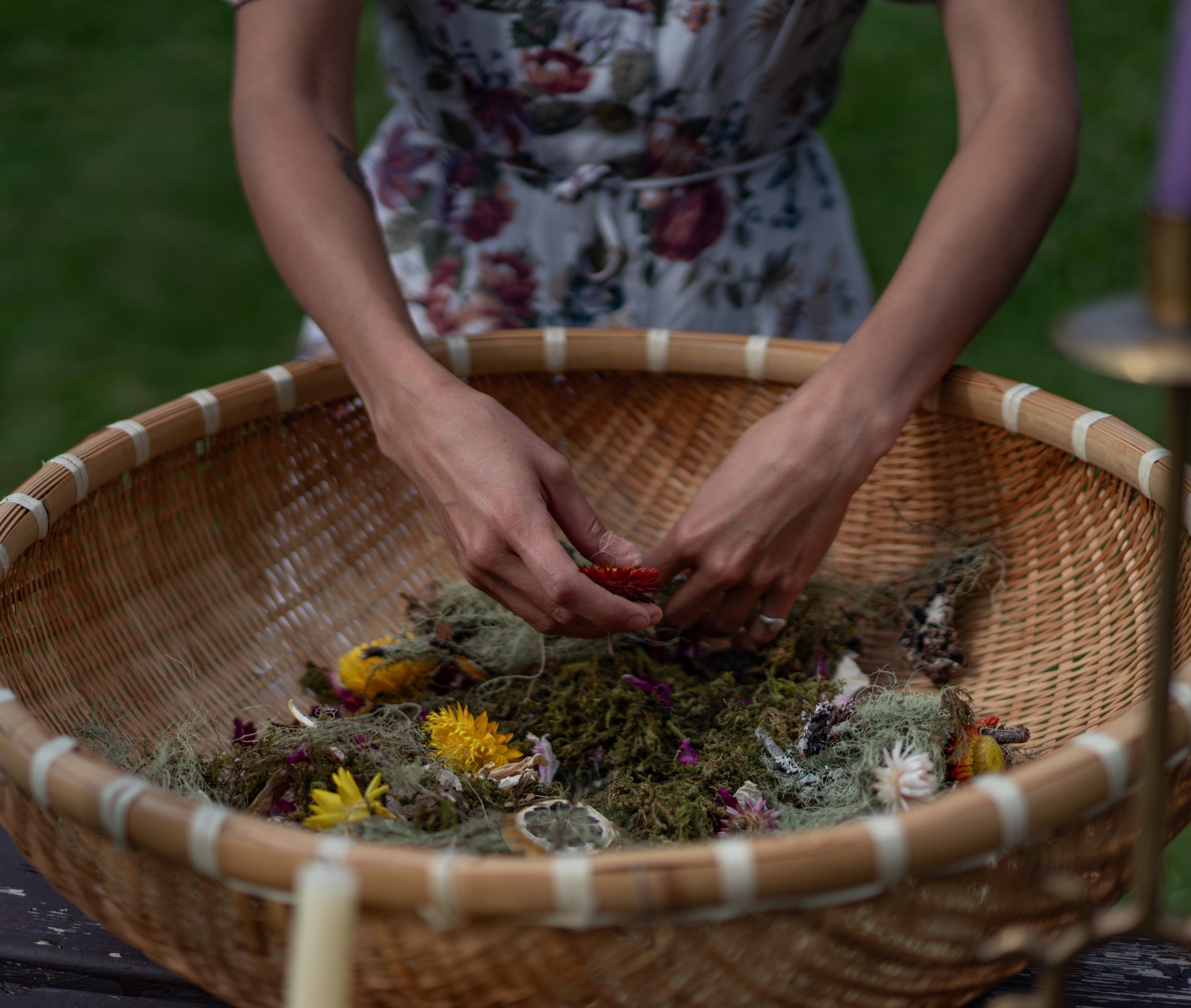 Hands arranging dried flowers and moss in woven basket centerpiece surrounded by candles