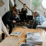People gathered around a wooden table with magazines on it, in a casual indoor setting.