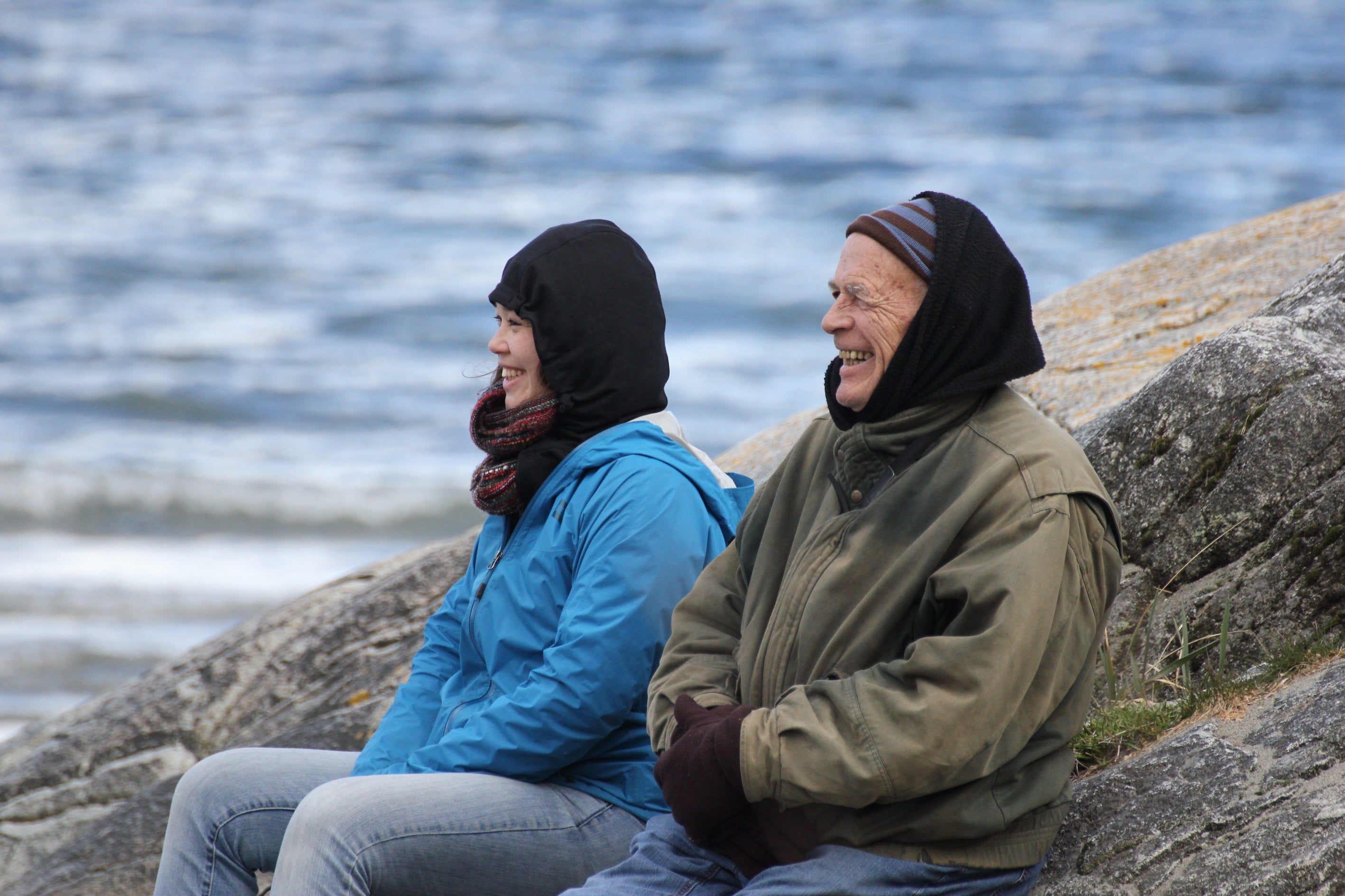 Two people sitting on rocks by a body of water, wearing winter clothing.