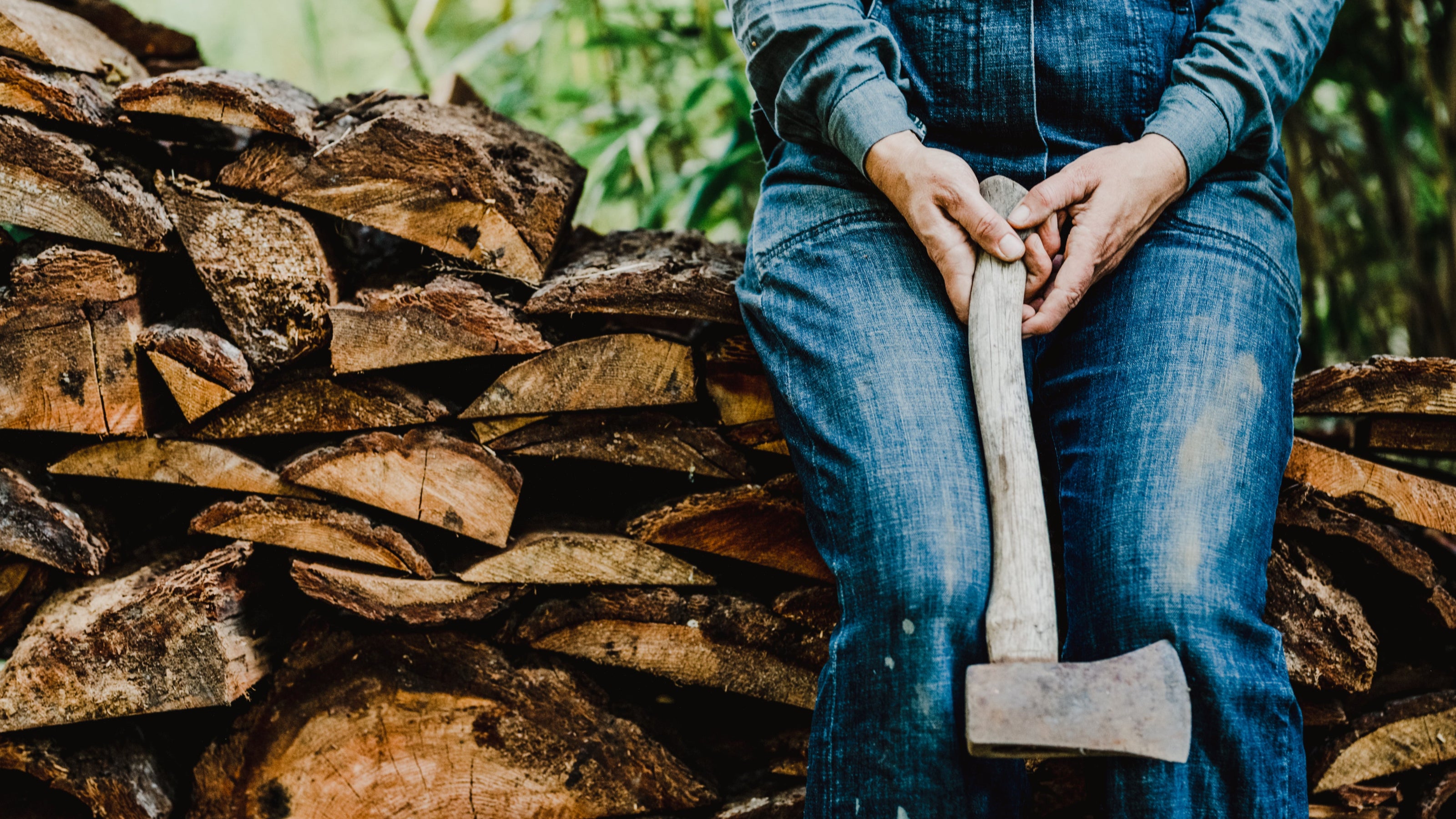 Person in blue overalls holding an axe next to a stack of firewood
