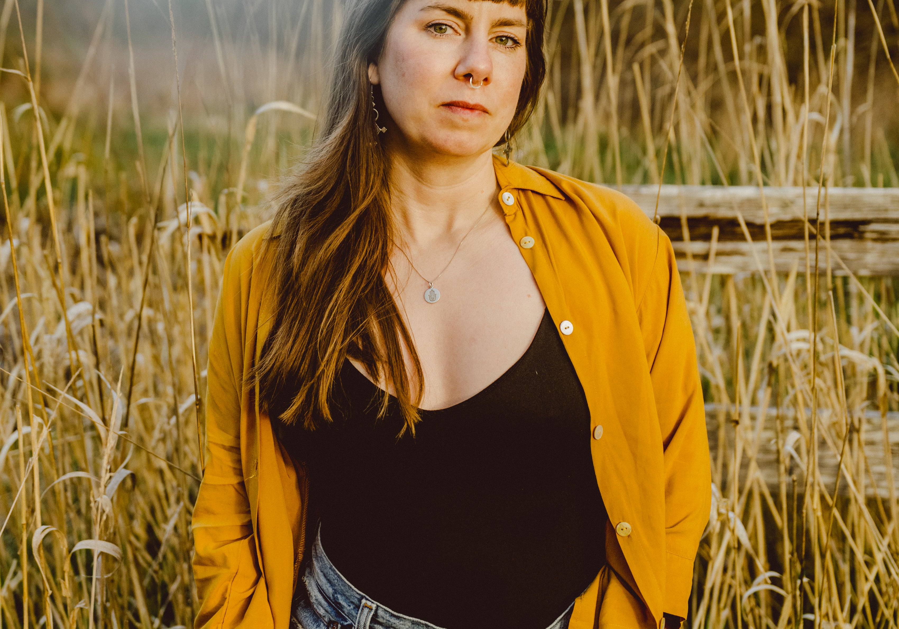 A woman standing in a field of tall dry grass, wearing a mustard yellow button-up shirt over a black top and blue jeans.