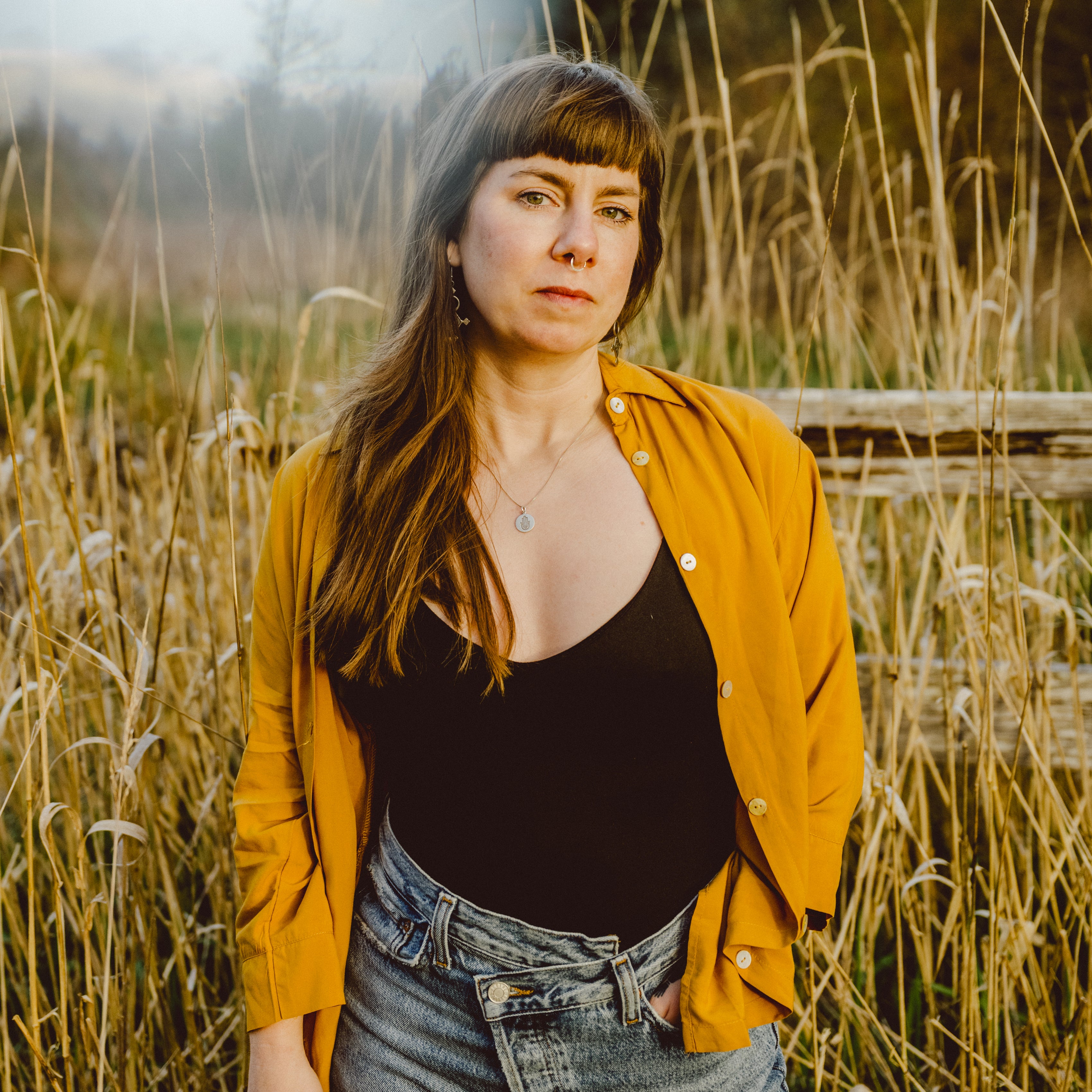 A woman standing in a field of tall dry grass, wearing a mustard yellow button-up shirt over a black top and blue jeans.