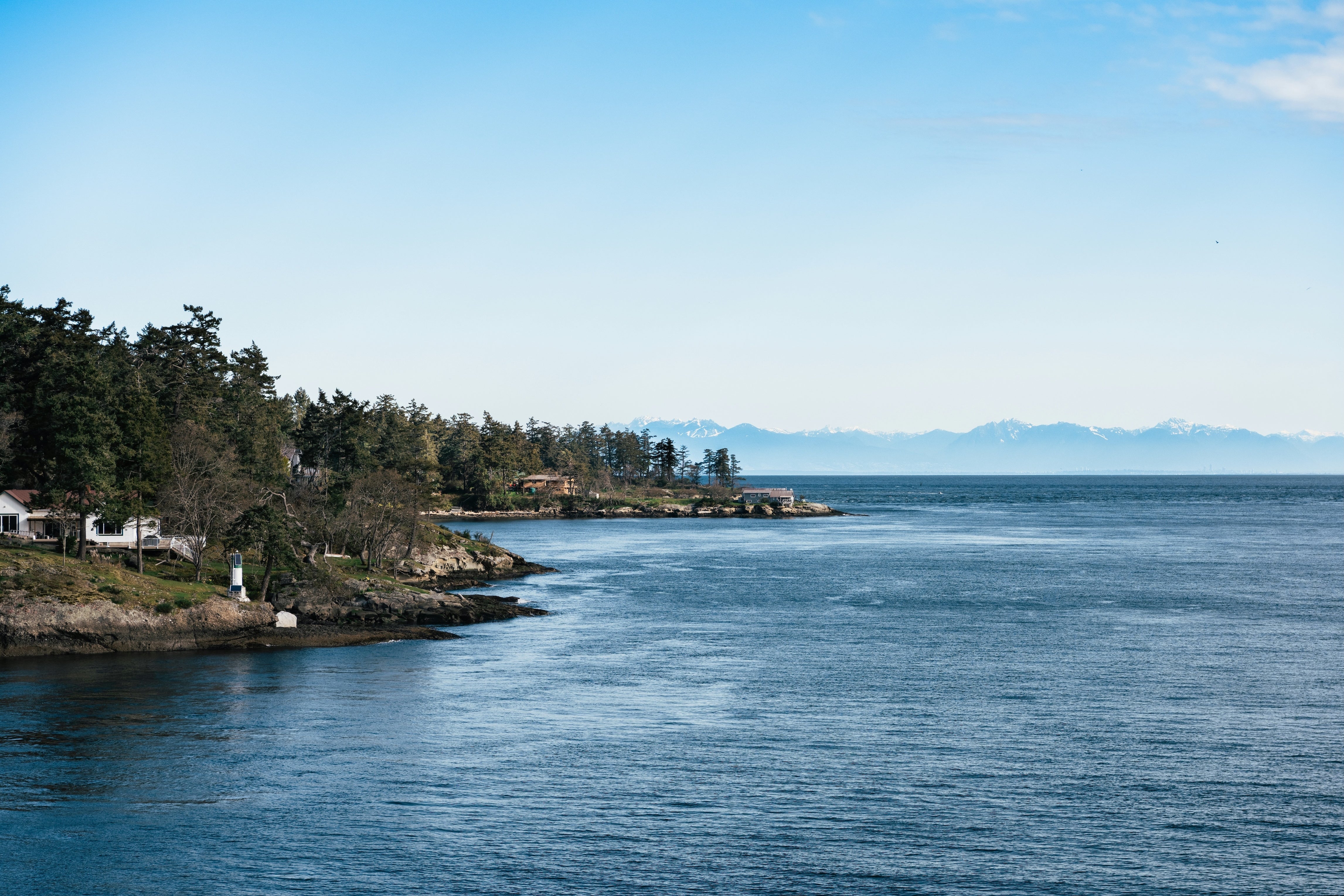 A scenic coastal landscape with deep blue water in the foreground, a rocky shoreline lined with dark green trees on the left, and distant mountains under a pale blue sky.