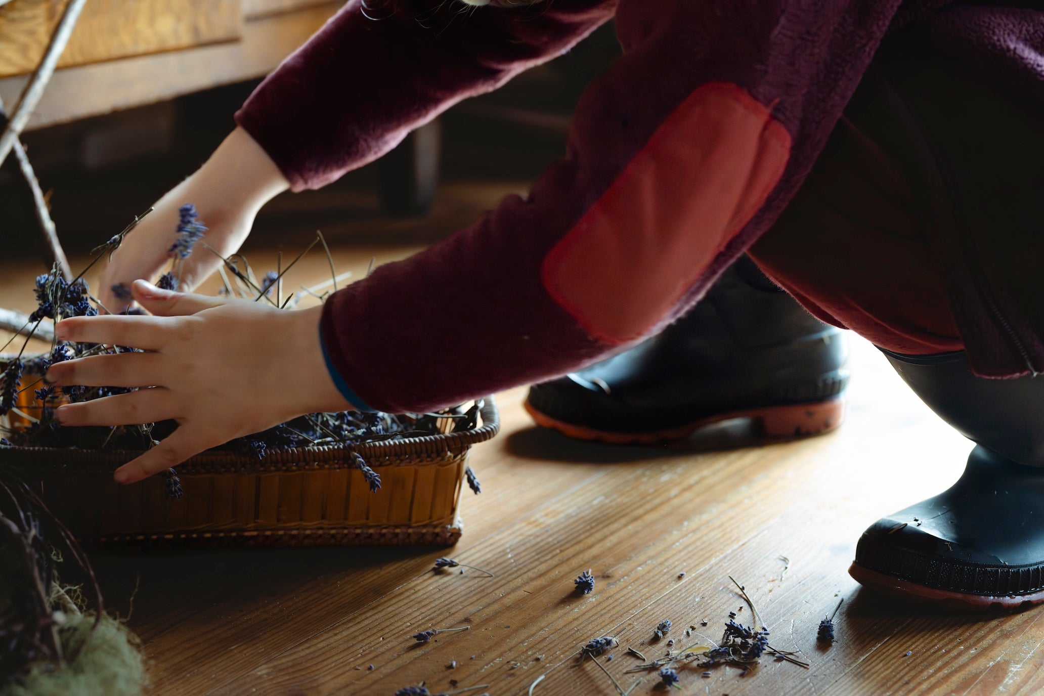 Person arranging dried plants in a woven basket on a wooden floor.