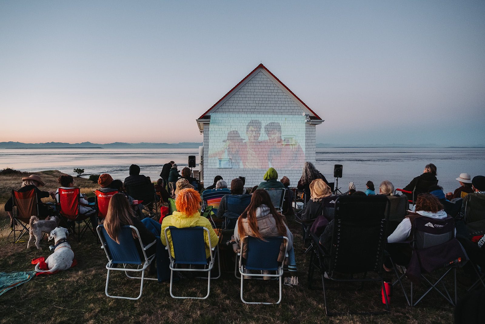 People watching a movie projected onto a small building by the sea at sunset.