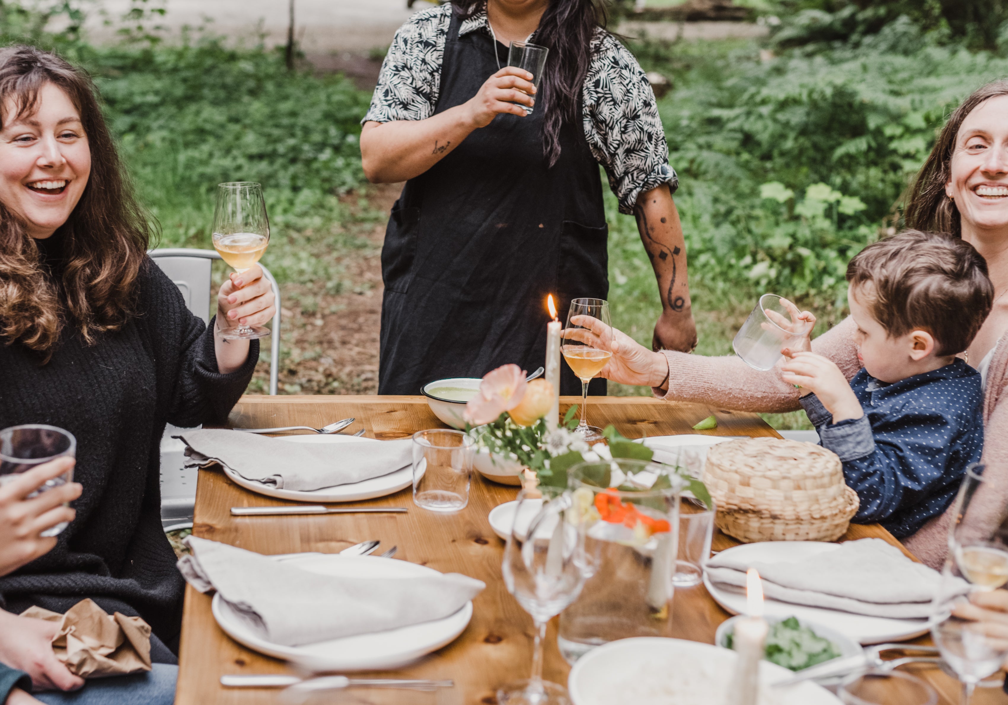 Group of people enjoying a meal outdoors at a wooden table with nature in the background.