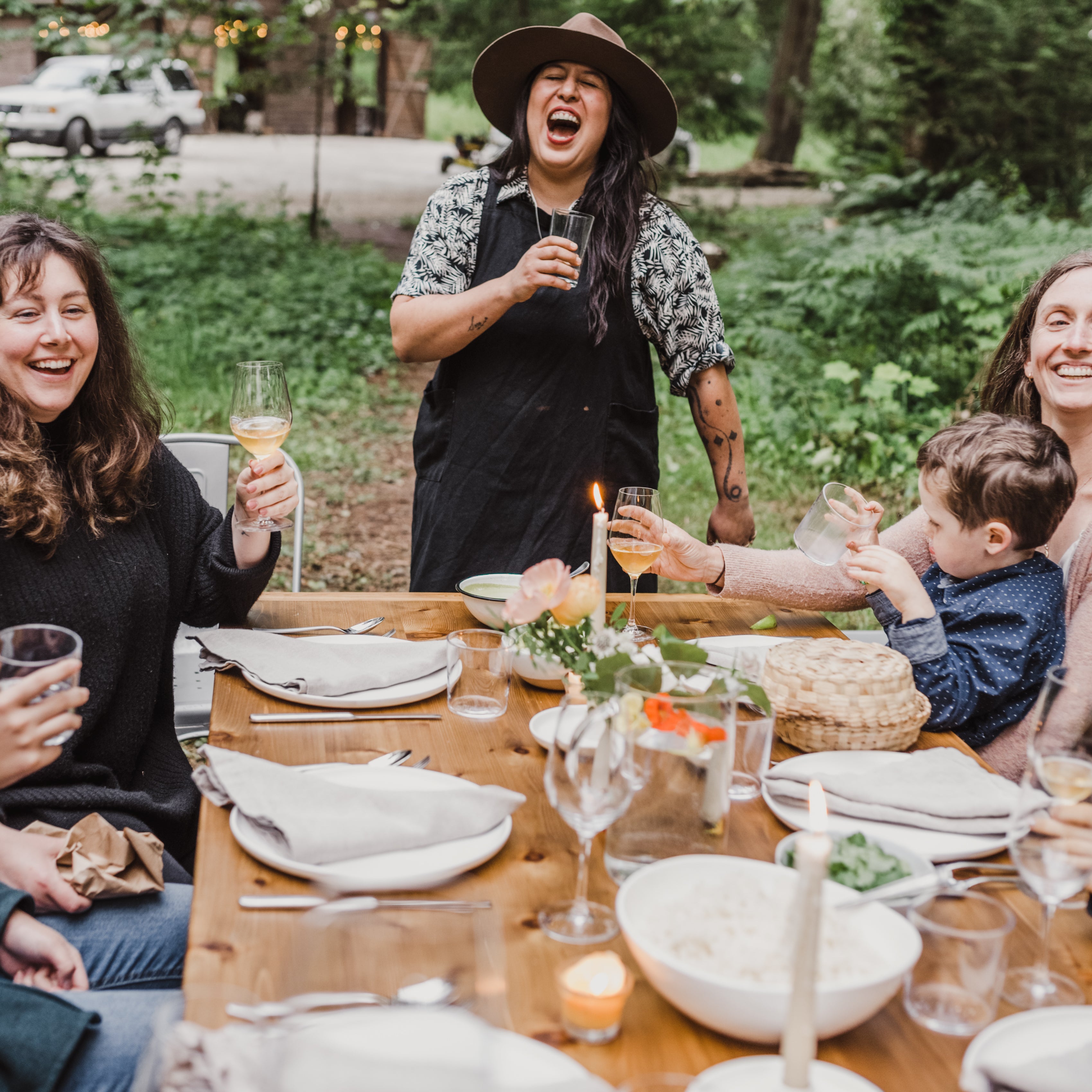Group of people enjoying a meal outdoors at a wooden table with nature in the background.