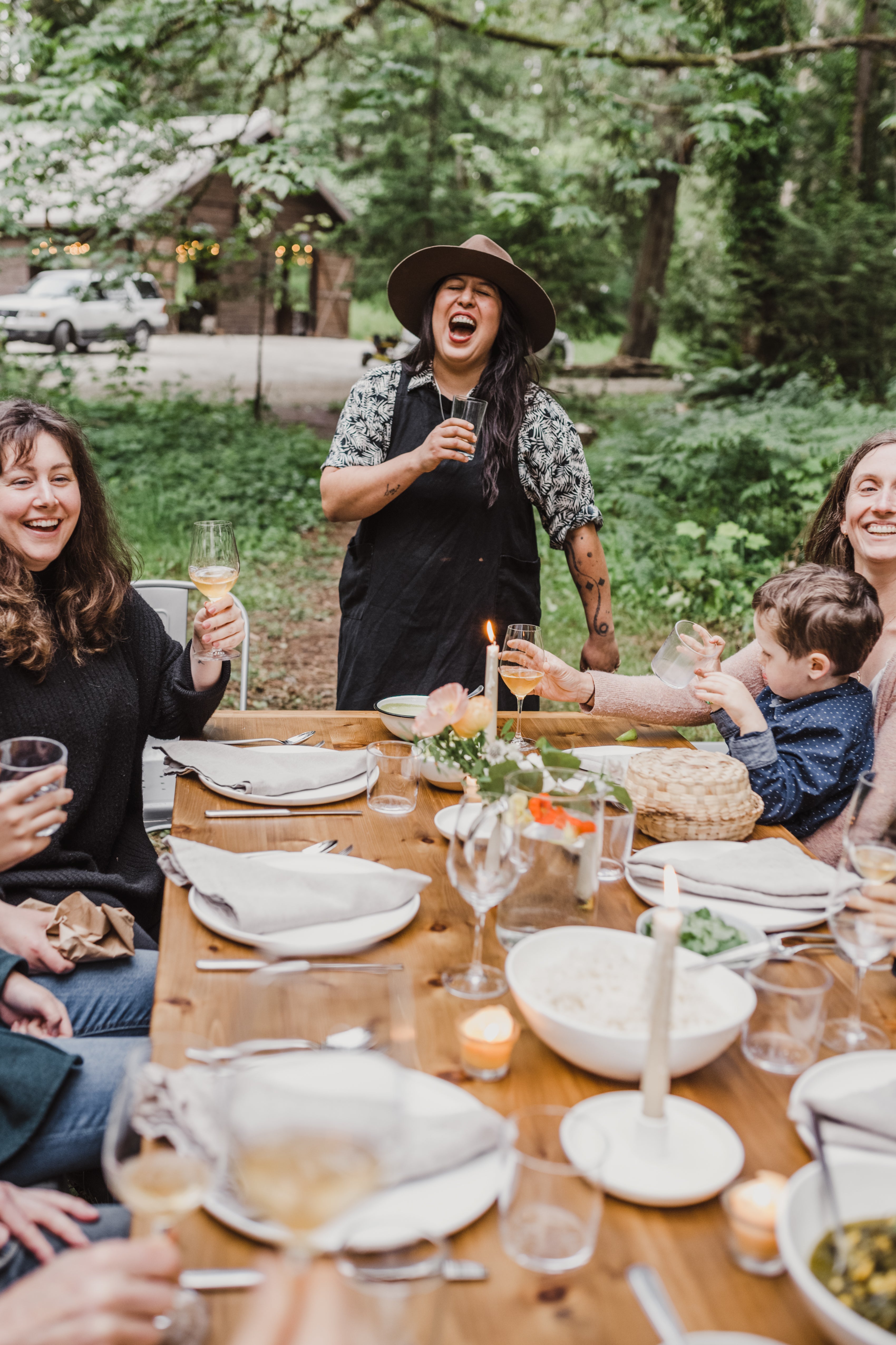Group of people enjoying a meal outdoors at a wooden table with nature in the background.
